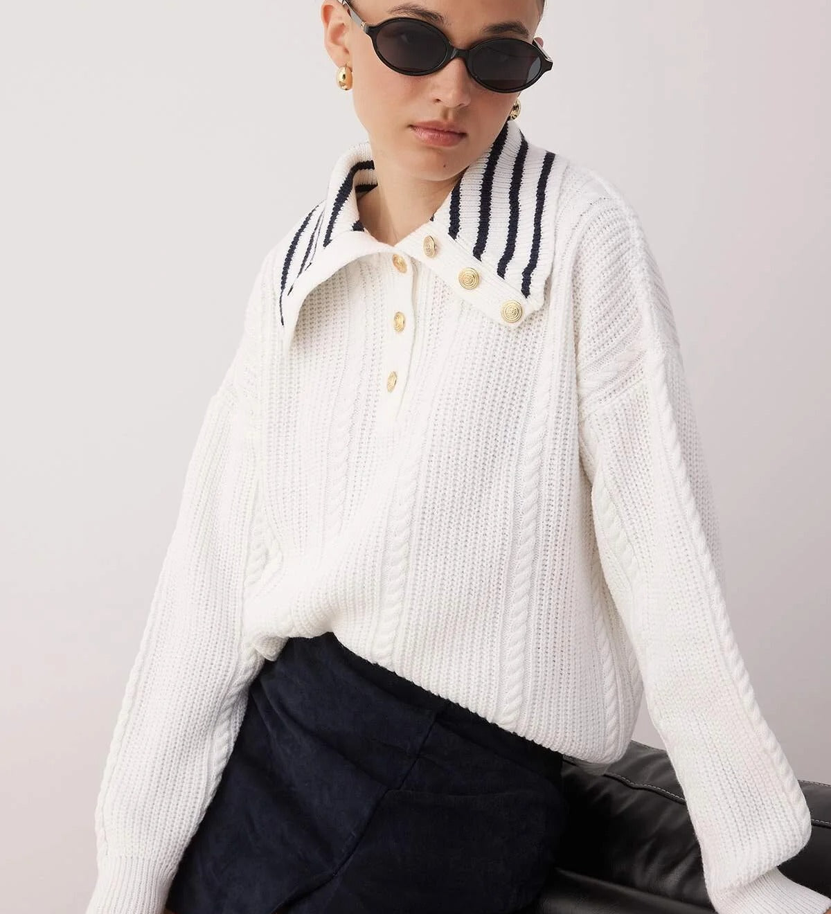 Woman wearing a white ribbed sweater with striped collar and black shorts against a plain background