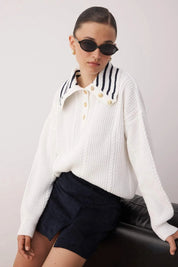 Woman wearing a white ribbed sweater with striped collar and black shorts against a plain background