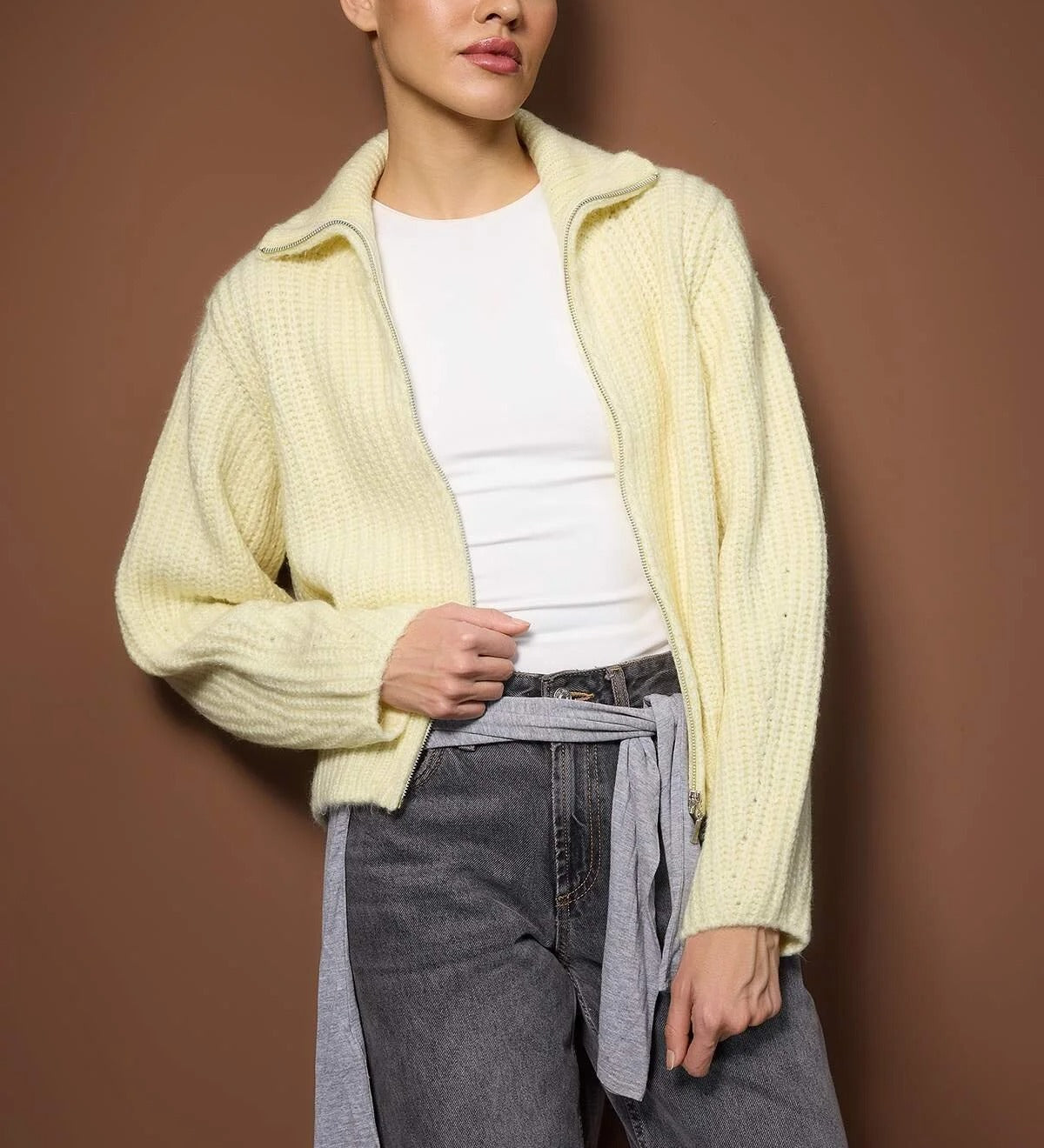 Woman wearing a light yellow cardigan over a white shirt with gray pants against a brown background
