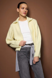 Woman wearing a light yellow cardigan over a white shirt with gray pants against a brown background