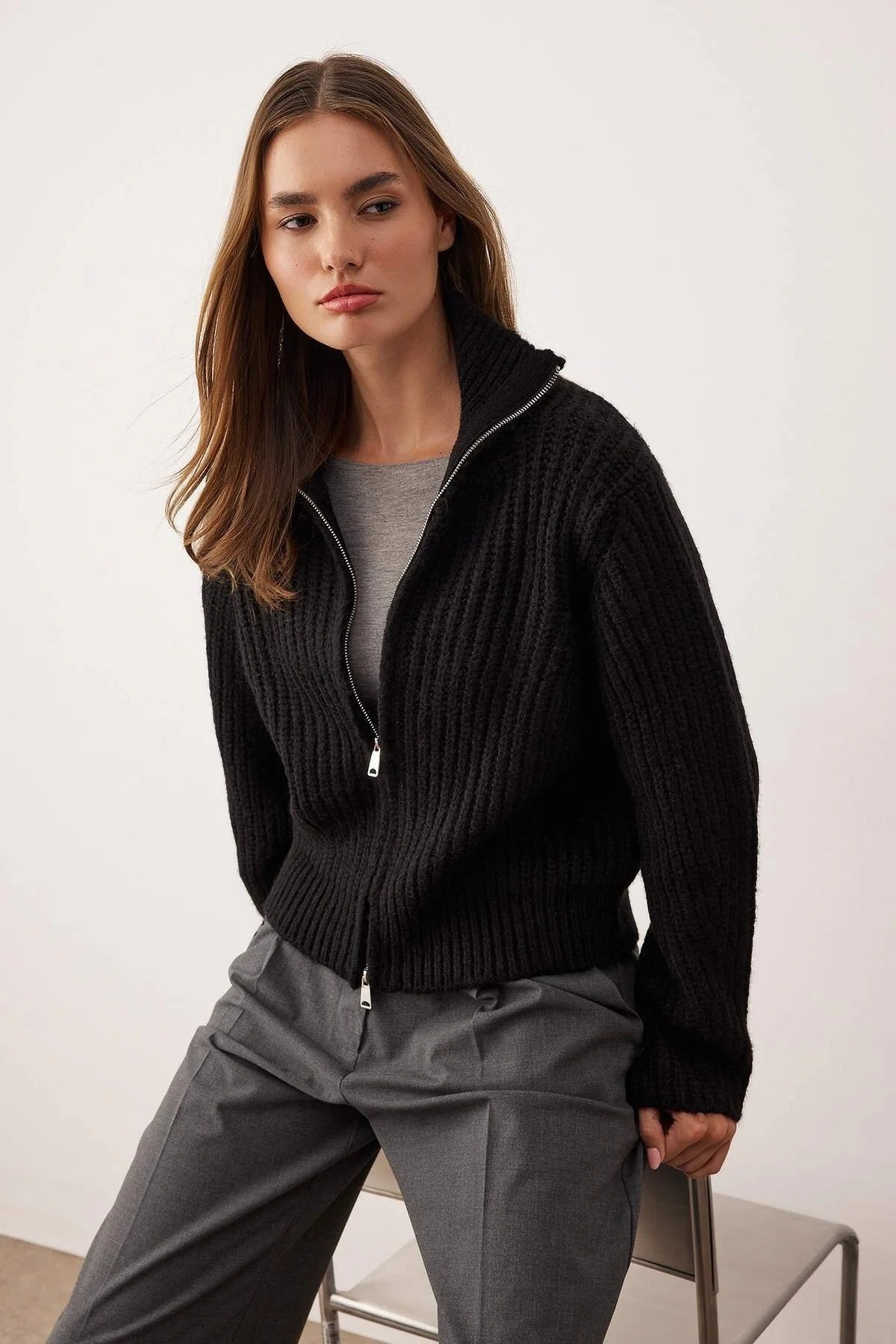 Woman wearing a black zip-up sweater over a gray shirt with gray pants on a white background