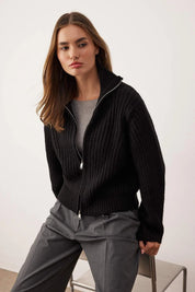 Woman wearing a black zip-up sweater over a gray shirt with gray pants on a white background