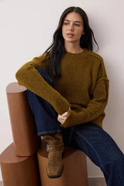Woman wearing a mustard yellow sweater and blue jeans sitting on wooden stools against a white wall.