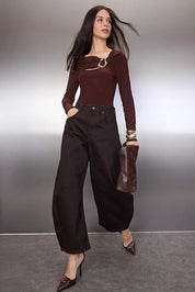 Woman in brown top and black pants holding a brown bag against a gray background