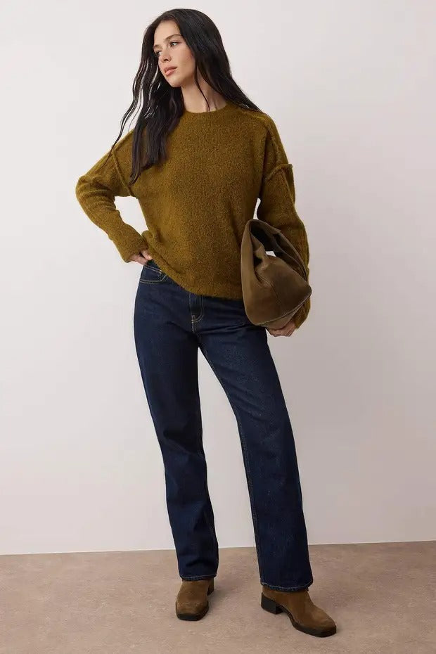 Woman wearing a mustard yellow sweater, dark blue jeans, and brown shoes against a white background