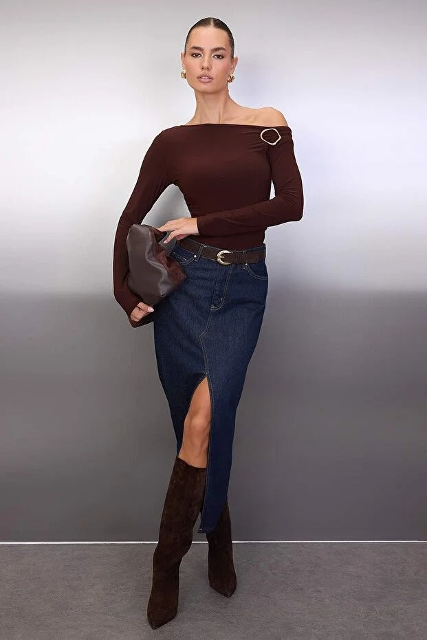 Woman wearing a brown off-shoulder top and blue jeans with a belt, standing against a gray background.