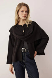 Woman wearing a black poncho over a gray top and blue jeans on a plain background