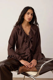 Woman wearing a brown ruffled blouse and pants sitting on a chair.