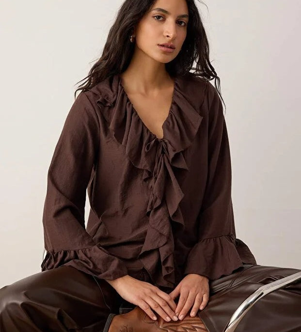 Woman wearing a brown ruffled blouse and pants sitting on a chair.