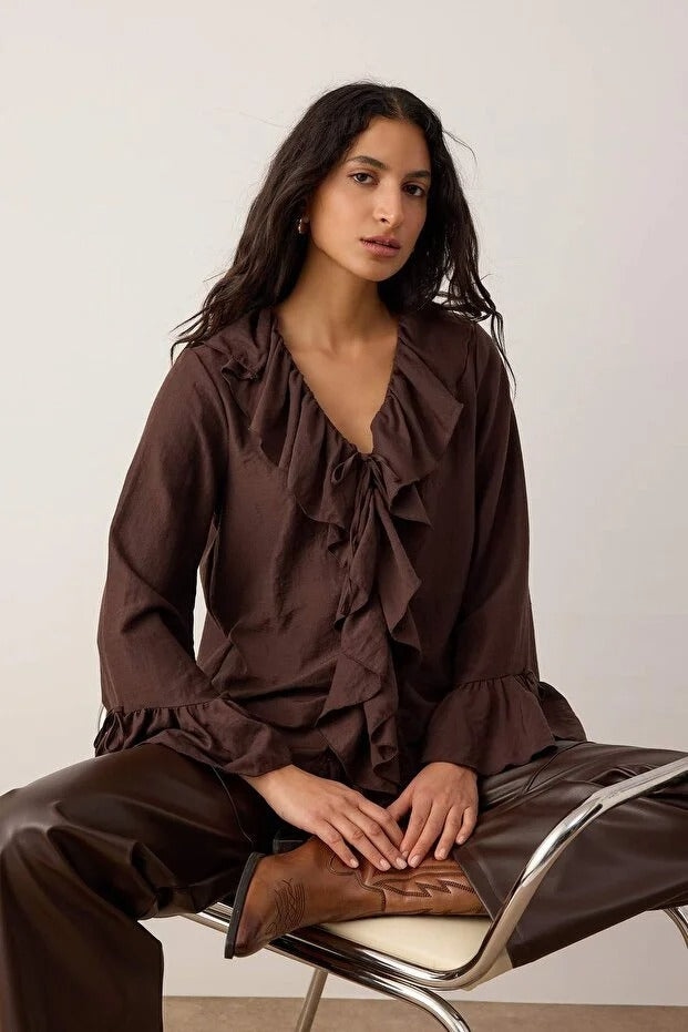 Woman wearing a brown ruffled blouse and pants sitting on a chair.