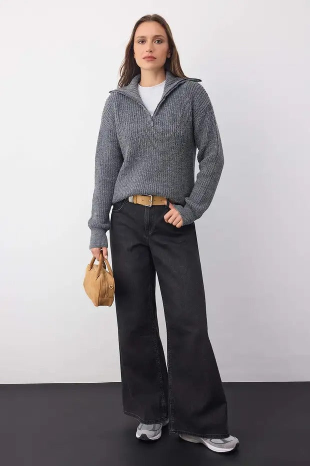 Woman wearing a gray sweater and black pants, holding a beige bag against a white background