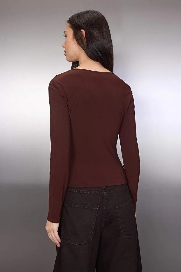 Woman wearing a brown long-sleeve top and dark pants against a plain background