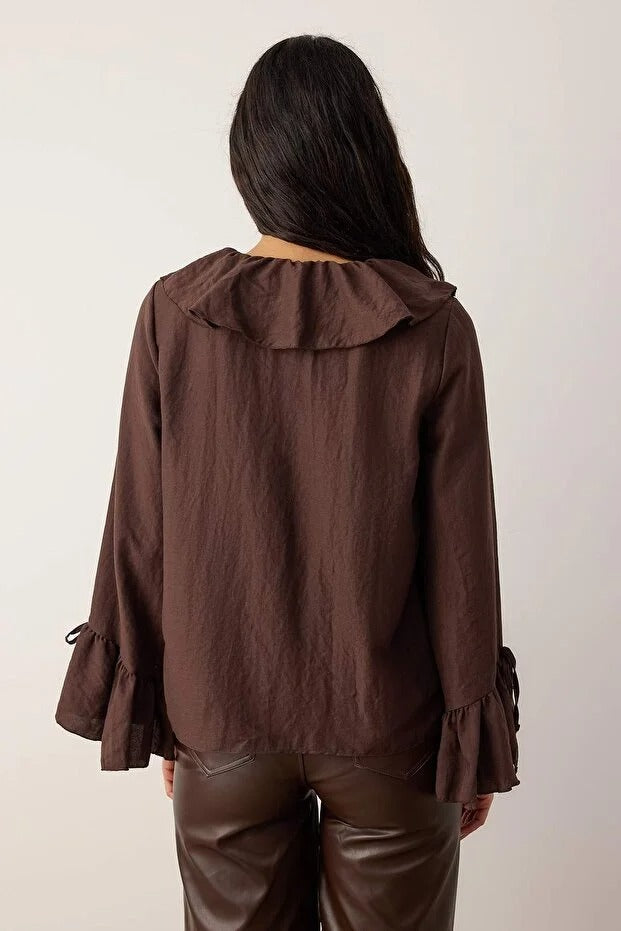 Person wearing a brown jacket with ruffled sleeves on a plain background