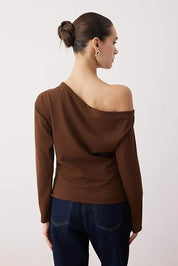 Woman wearing a brown one-shoulder top and dark jeans on a light pink background